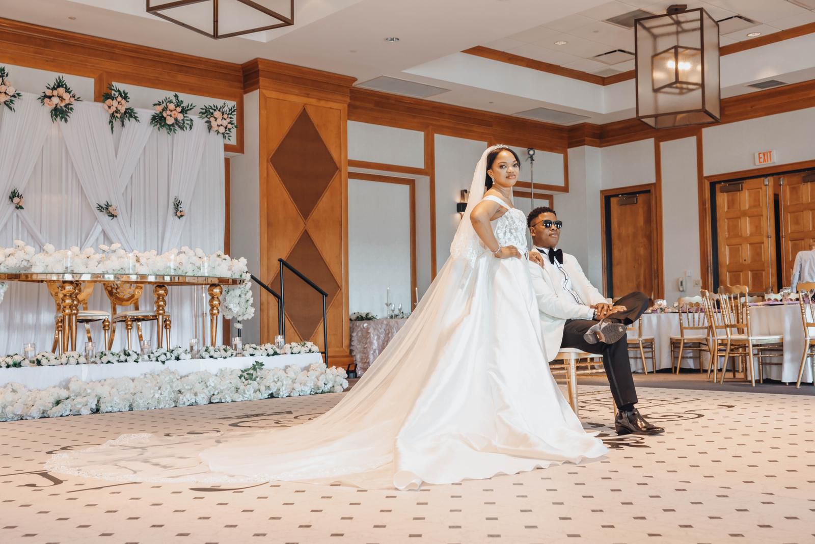 Bride with a long cathedral train beside her seated groom in the ballroom